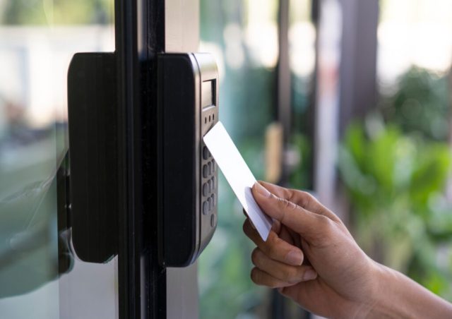 A hand holds a white key card near a black card reader on a glass door, suggesting entry access. Background shows blurred greenery.